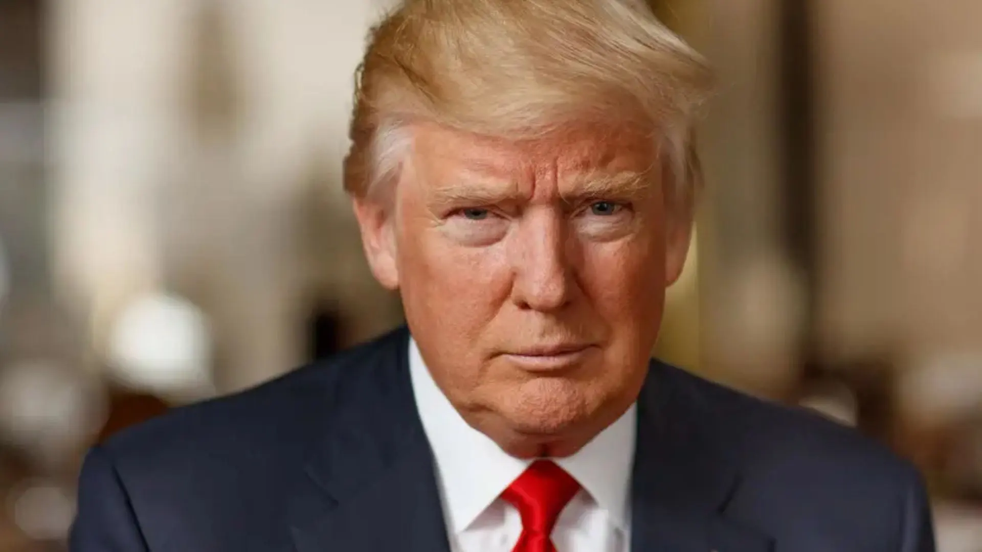 The image shows a man with a serious and expressive face, with blond hair combed to the side and dressed in a dark suit, white shirt, and red tie. His gaze is direct, and his expression conveys determination or concentration. The background is slightly blurred, helping to focus attention on the face and details of the main figure. The photograph appears to be taken in a formal or professional setting, possibly in a government building or high-profile context.